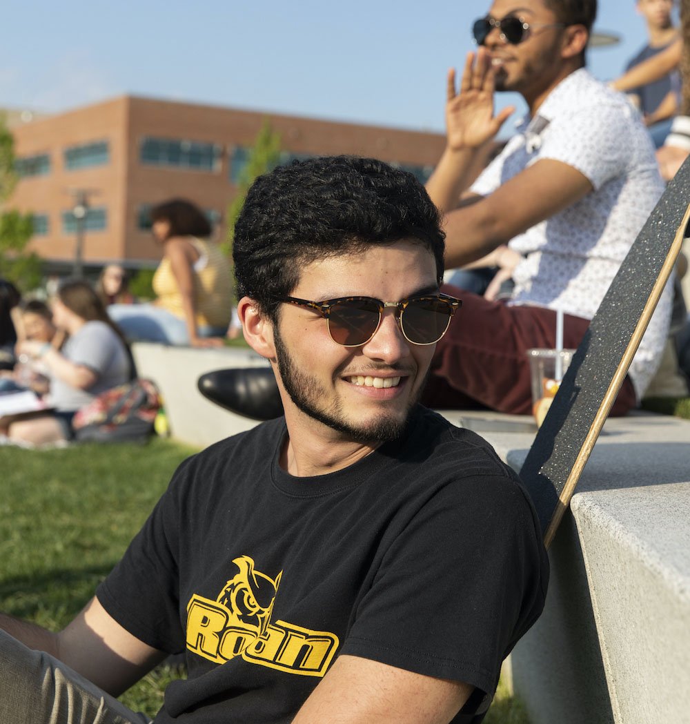 Male student sits outside, smiling off to the distance while wearing sunglasses.